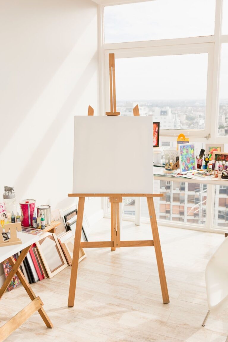 A wooden H-frame easel with a blank canvas in a studio by the window